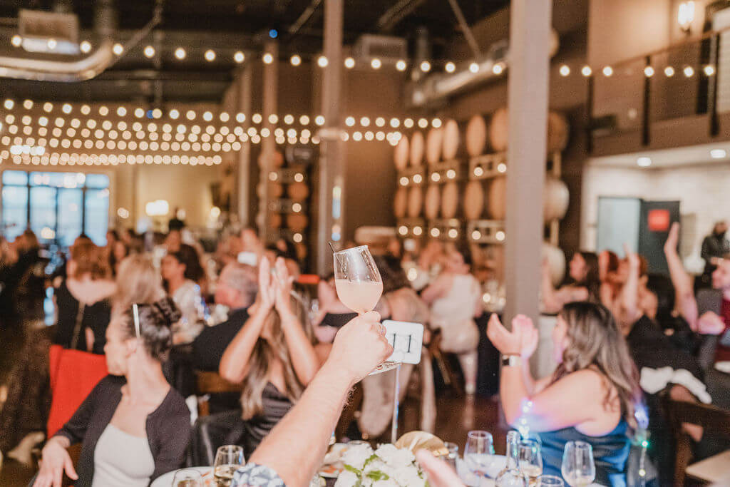 A private event at Domenico Winery San Carlos with a wine glass raised at in the main Barrel room. Cekebration venue decorated with ceiling lights and people enjoying themselves at the event.