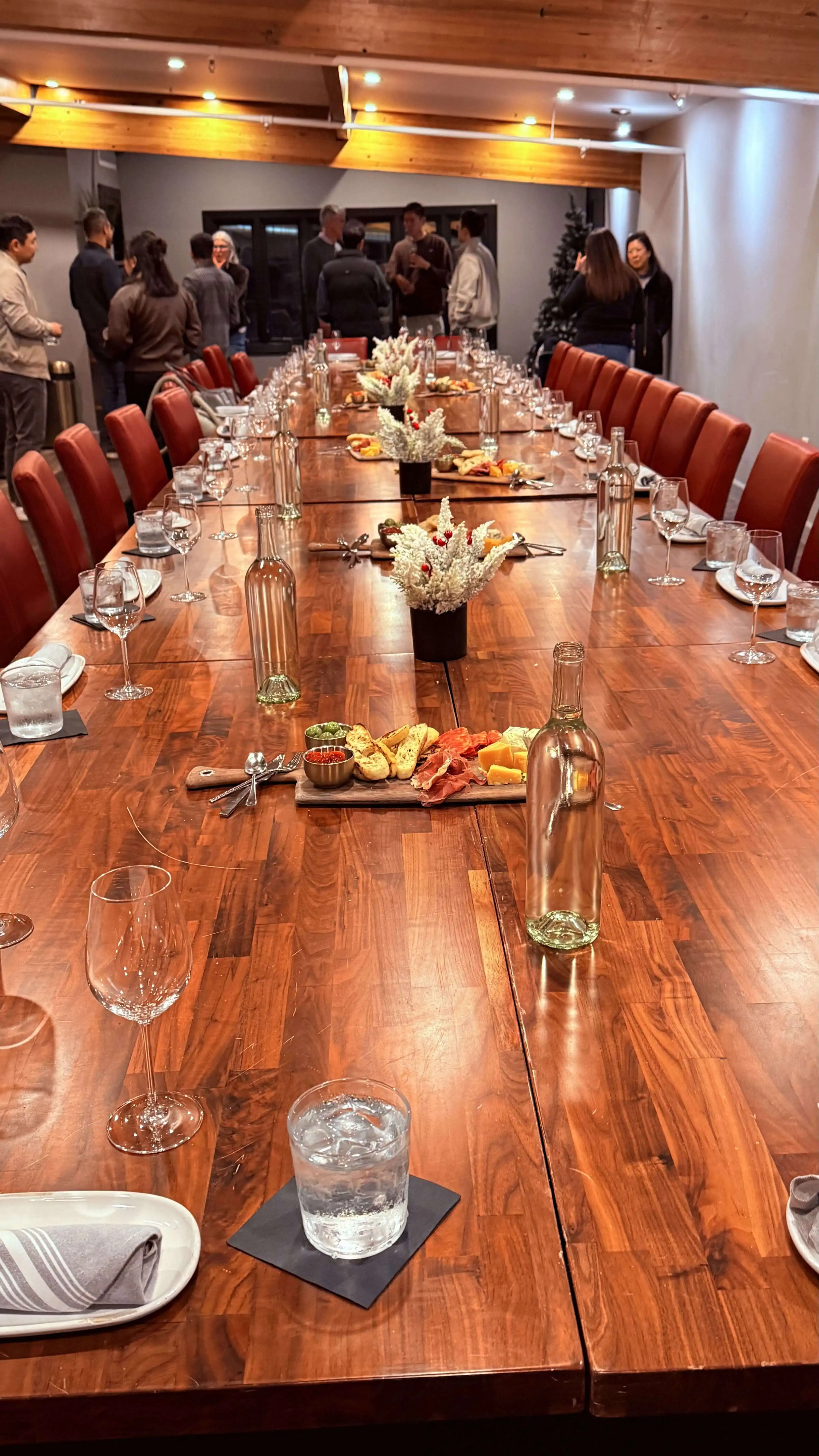 Long wooden dining table set for a group event with wine glasses, small floral centerpieces, and platters of appetizers, with guests mingling in the background.