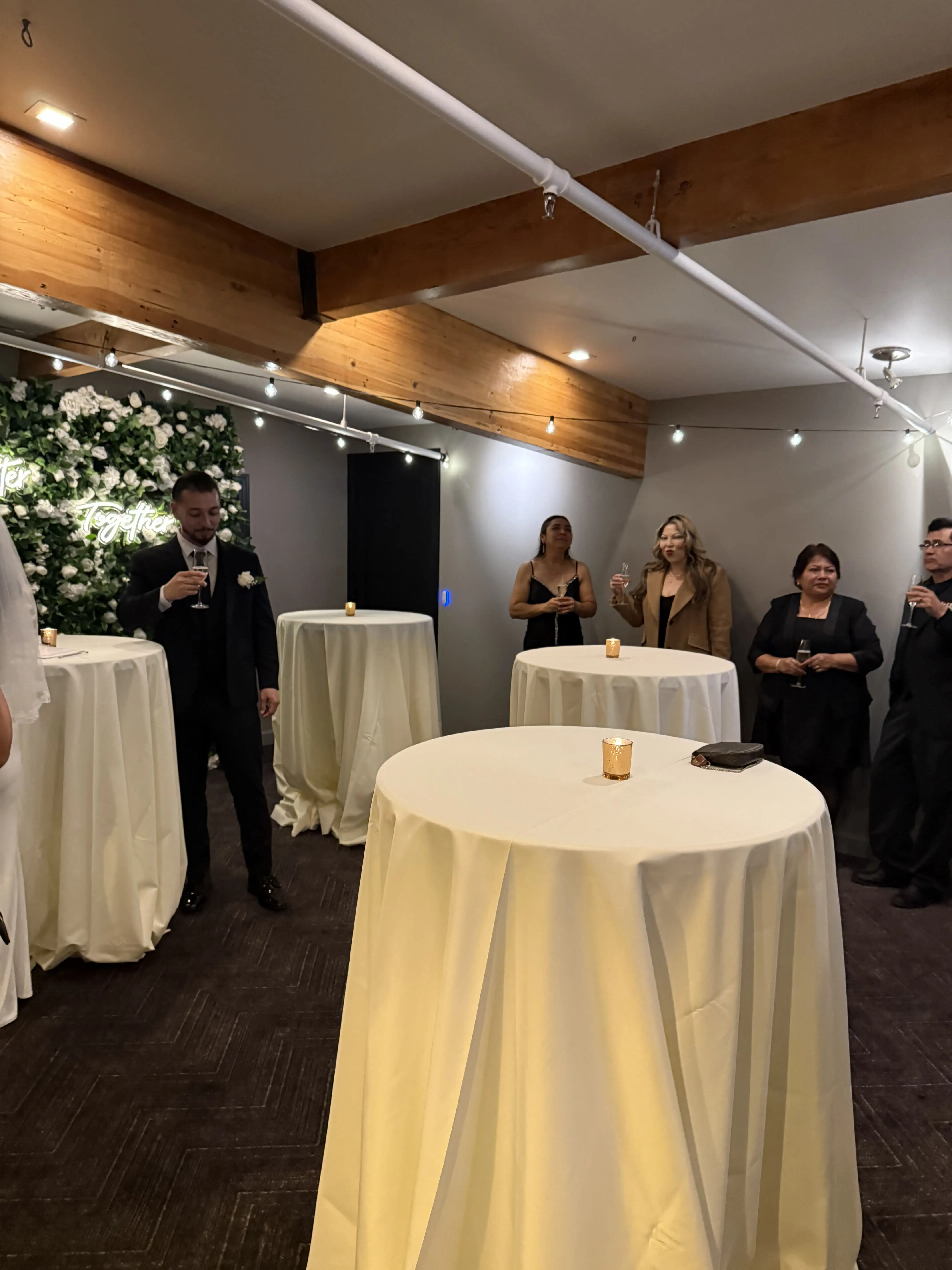 Wedding guests standing around tall cocktail tables, holding glasses for a toast in a decorated indoor reception area.