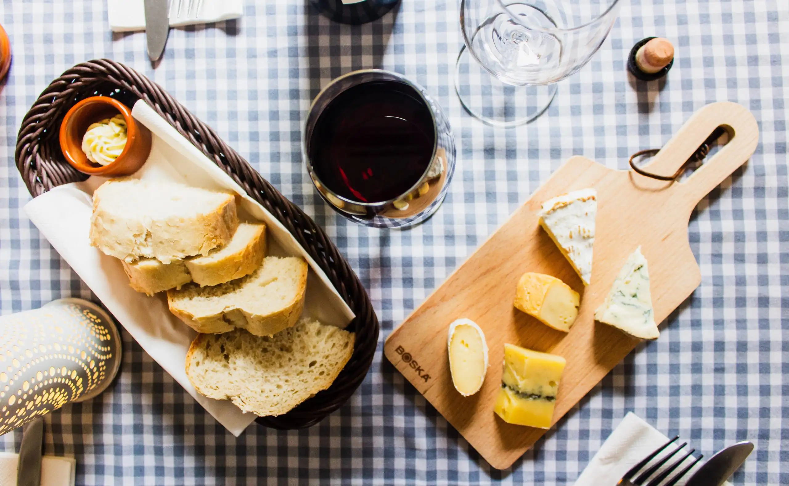 Fresh bread, a glass of red wine, and a wooden cheese platter on a checkered tablecloth