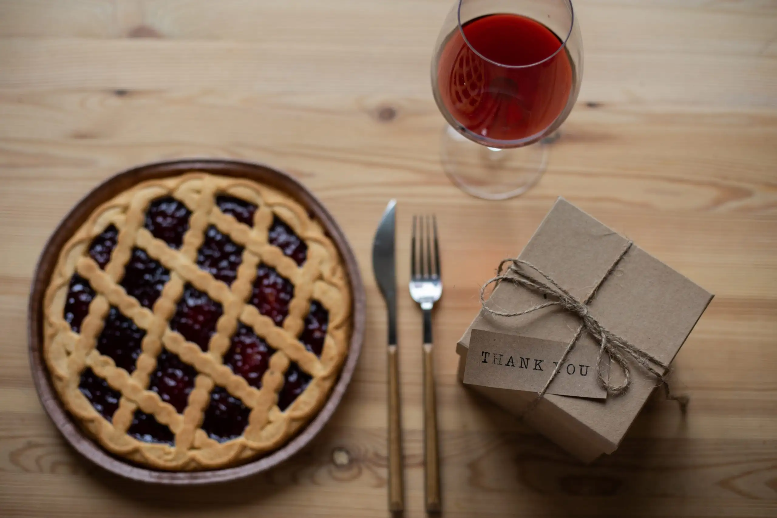 A wooden table setting featuring a lattice-top cherry pie, a glass of red wine, and a brown kraft paper gift box with a "Thank You" tag.