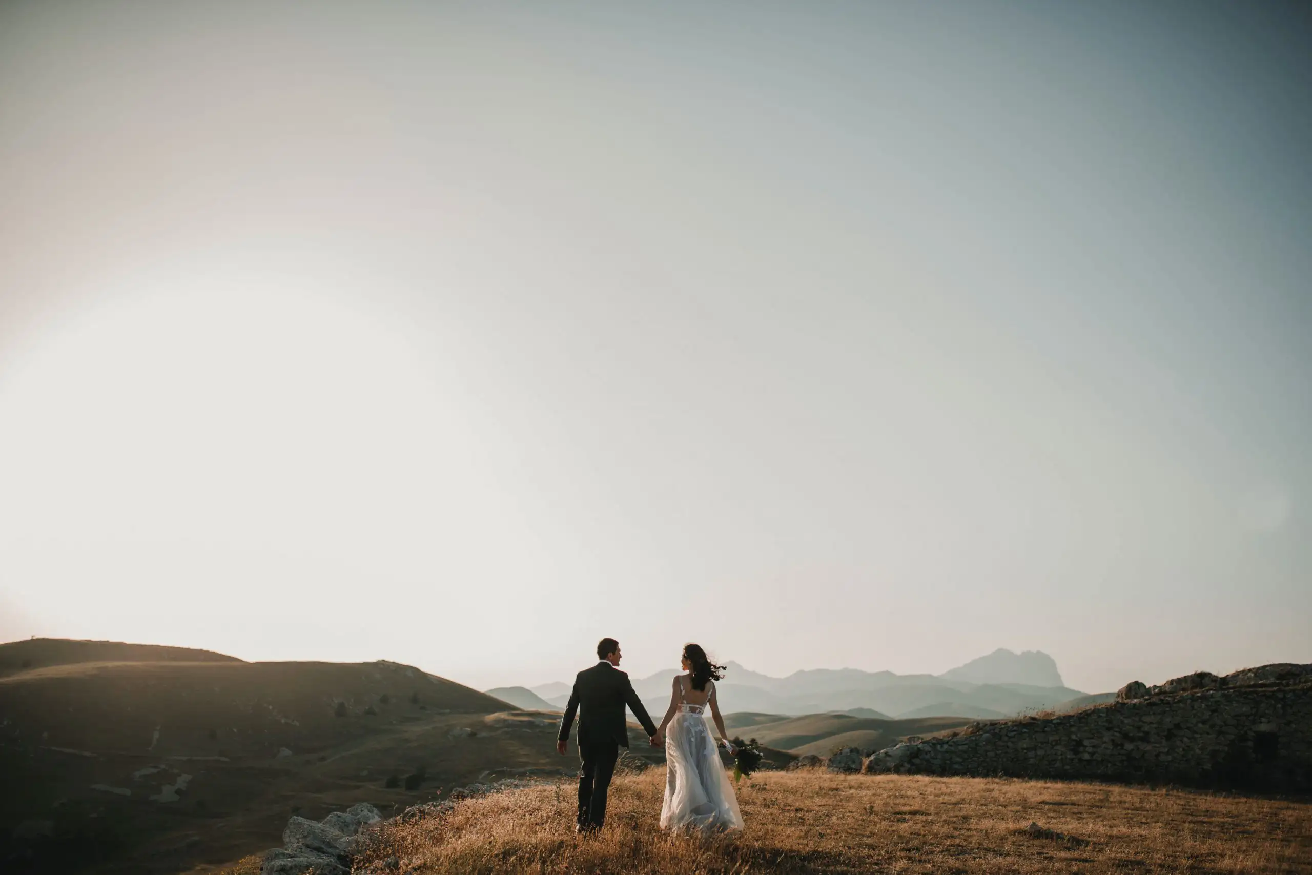 A couple in formal attire holding hands and walking across a grassy hilltop during golden hour with a vast mountain range in the distance.