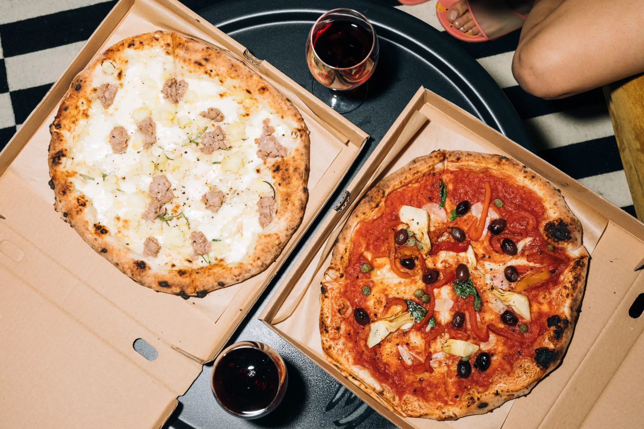 Two pizzas with different toppings served with red wine on a black table. One pizza has white sauce and sausage, and the other has tomato sauce, olives, and bell peppers.