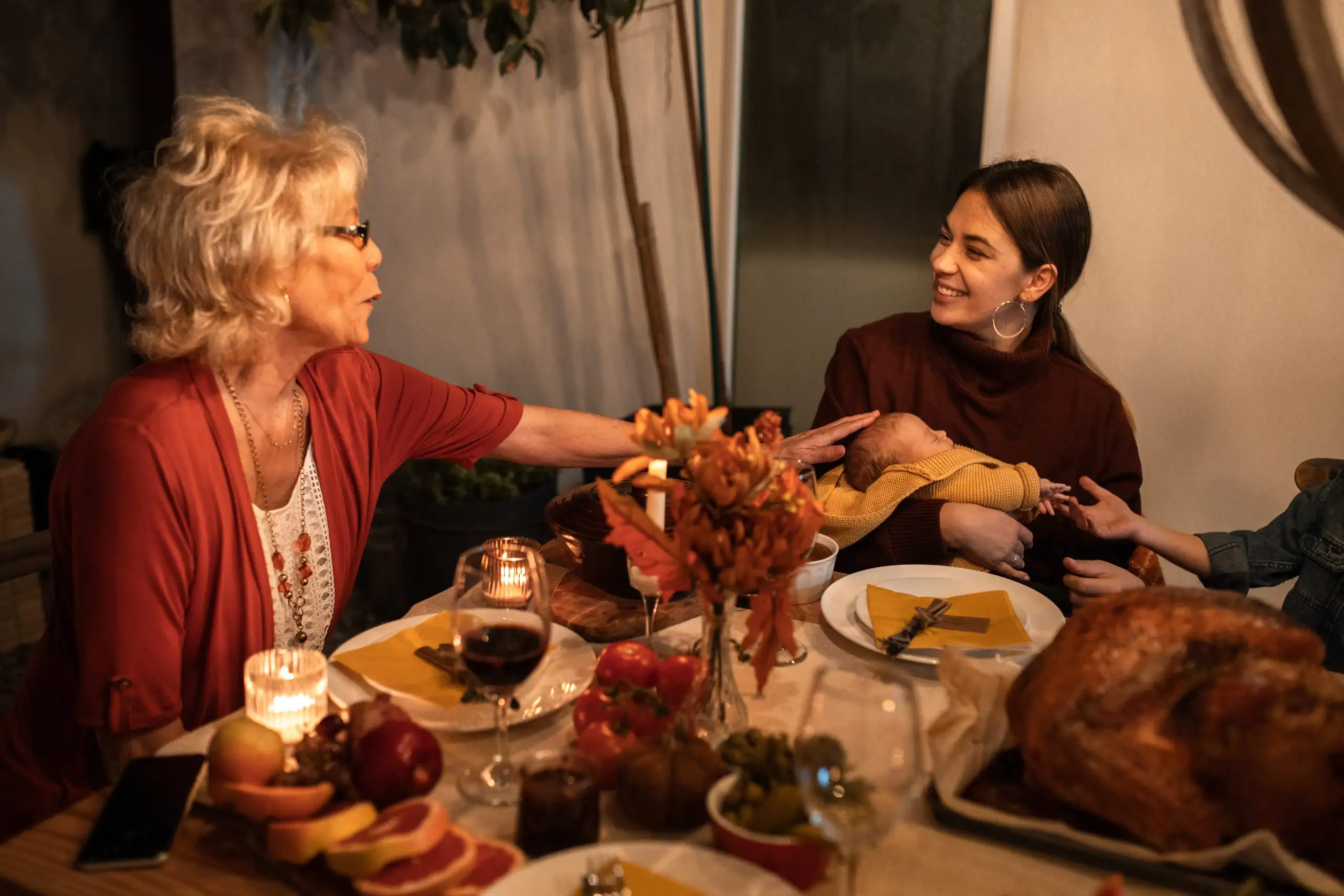 Grandmother talking with mother holding newborn baby at Thanksgiving dinner table with roasted turkey and candles
