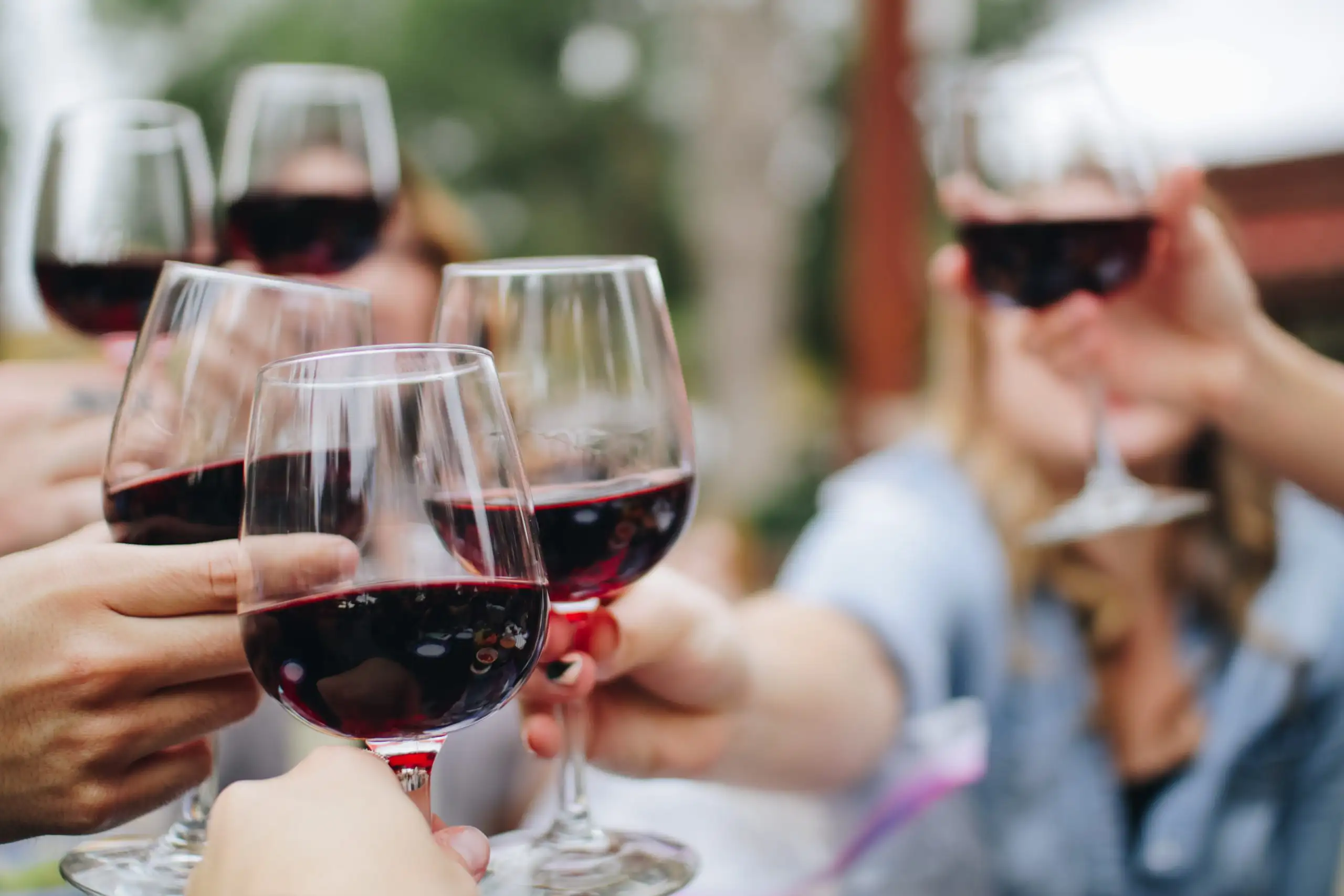 A group of friends holding up glasses of red wine for a toast in a blurred outdoor setting.