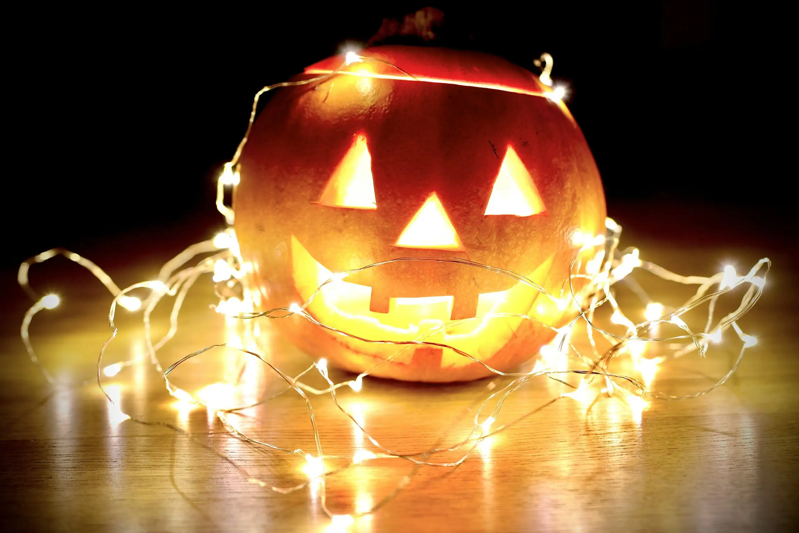 Carved Halloween jack-o’-lantern pumpkin glowing with candlelight and wrapped in warm white string lights on a wooden table against a dark background.