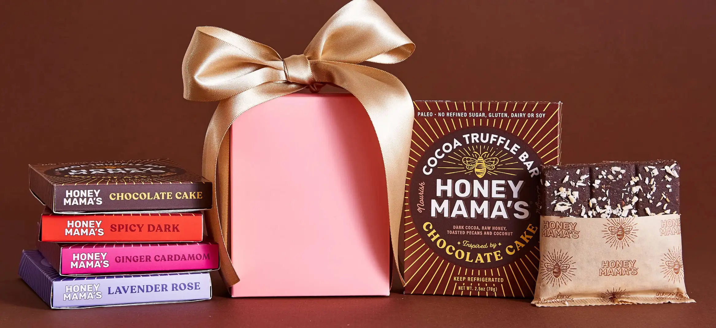 A collection of Honey Mama's cocoa truffle bars in various flavors like Lavender Rose and Spicy Dark, next to a pink gift box with a gold ribbon.