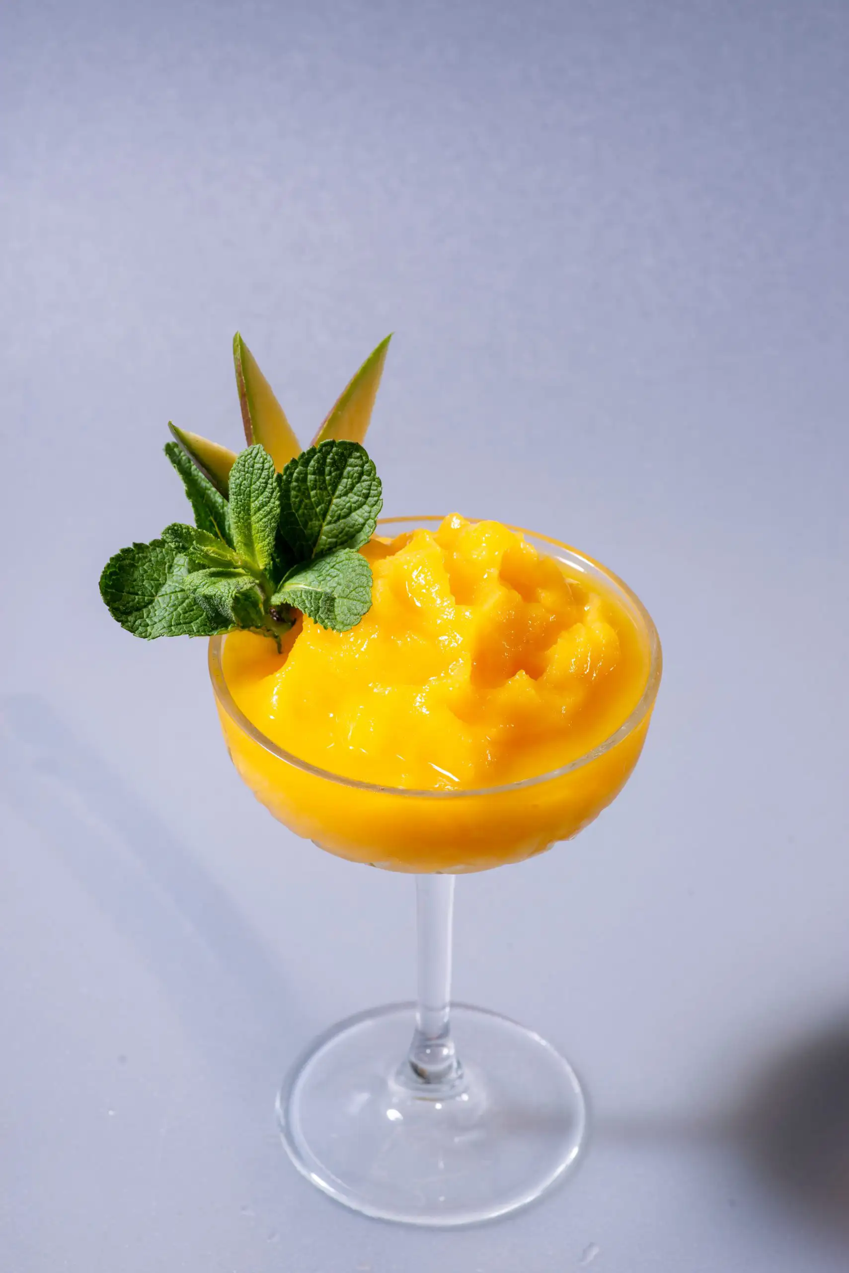 Vibrant yellow mango sorbet in a crystal coupe glass, garnished with fresh mint leaves and a thin fruit slice against a grey background.