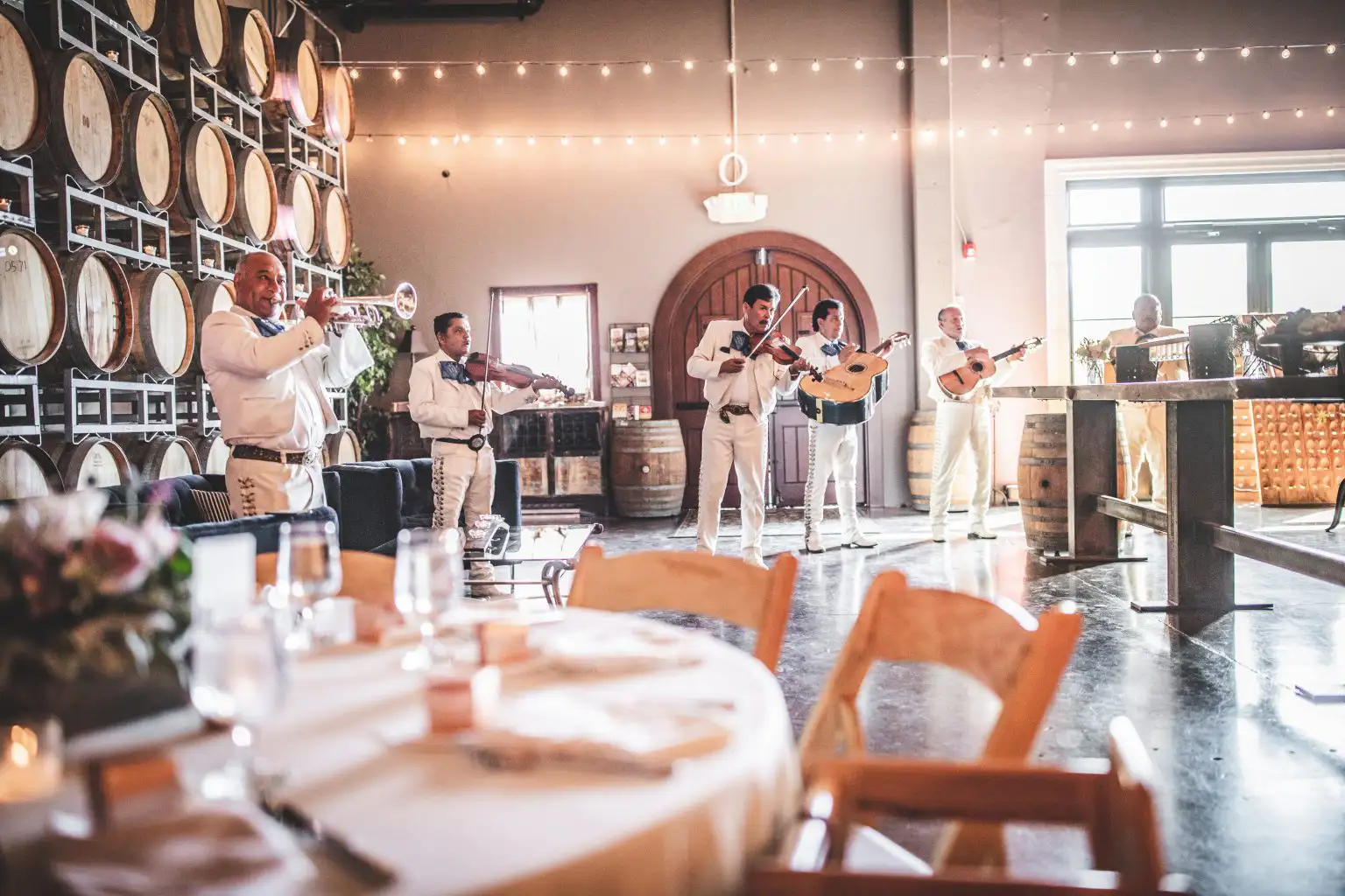 A five-piece mariachi band in white traditional suits playing instruments in a large room featuring a wall of stacked wooden wine barrels and string lights.