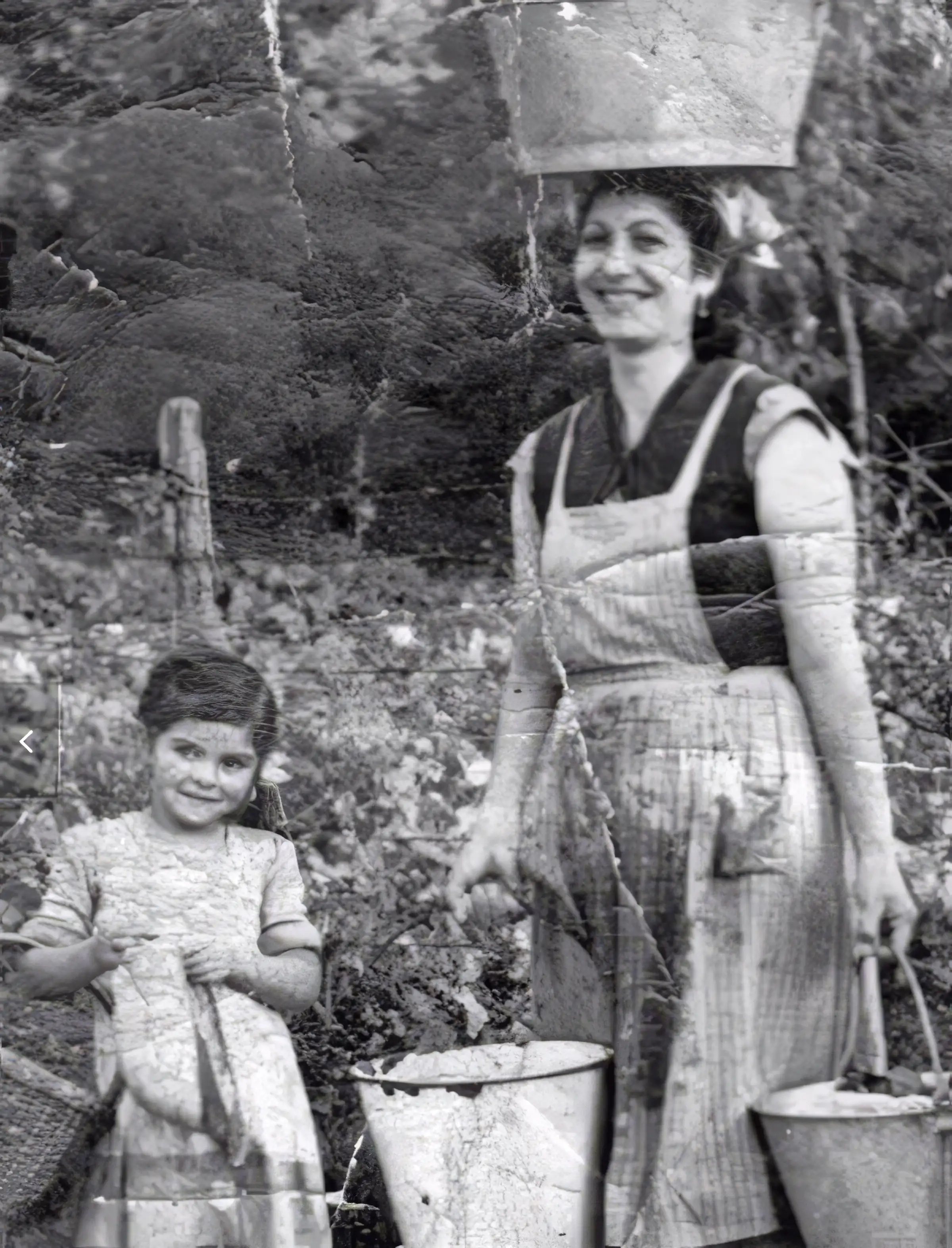Woman carrying metal water buckets and balancing a basin on her head while standing beside a young child in a rural field, vintage black-and-white photo.