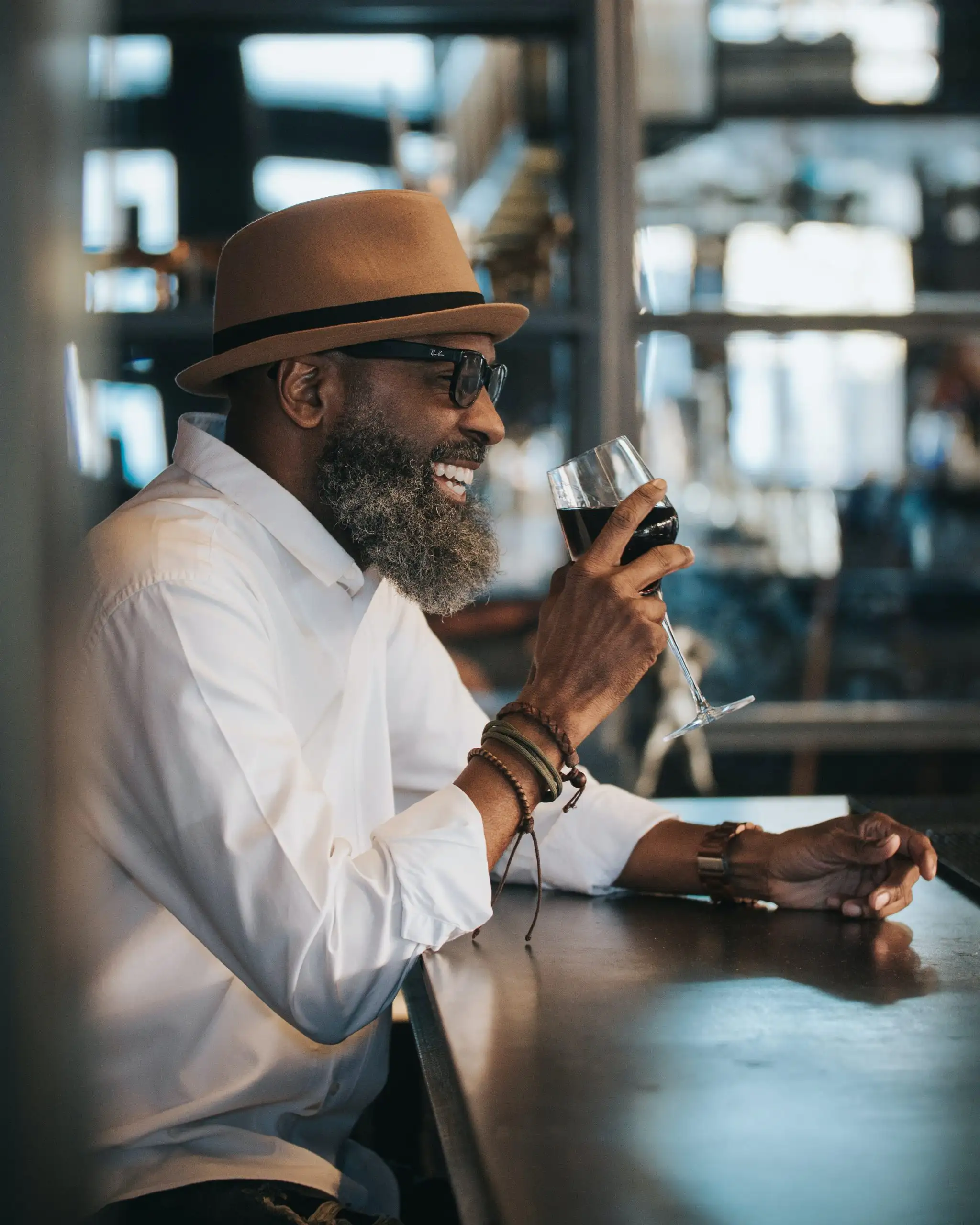 A smiling man in a beige hat, glasses, and white shirt sipping wine at a bar.
