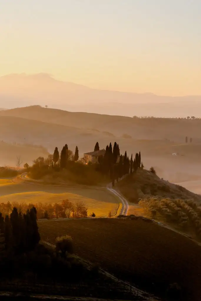 Sunrise over rolling hills in Tuscany with a farmhouse surrounded by cypress trees and a winding dirt road