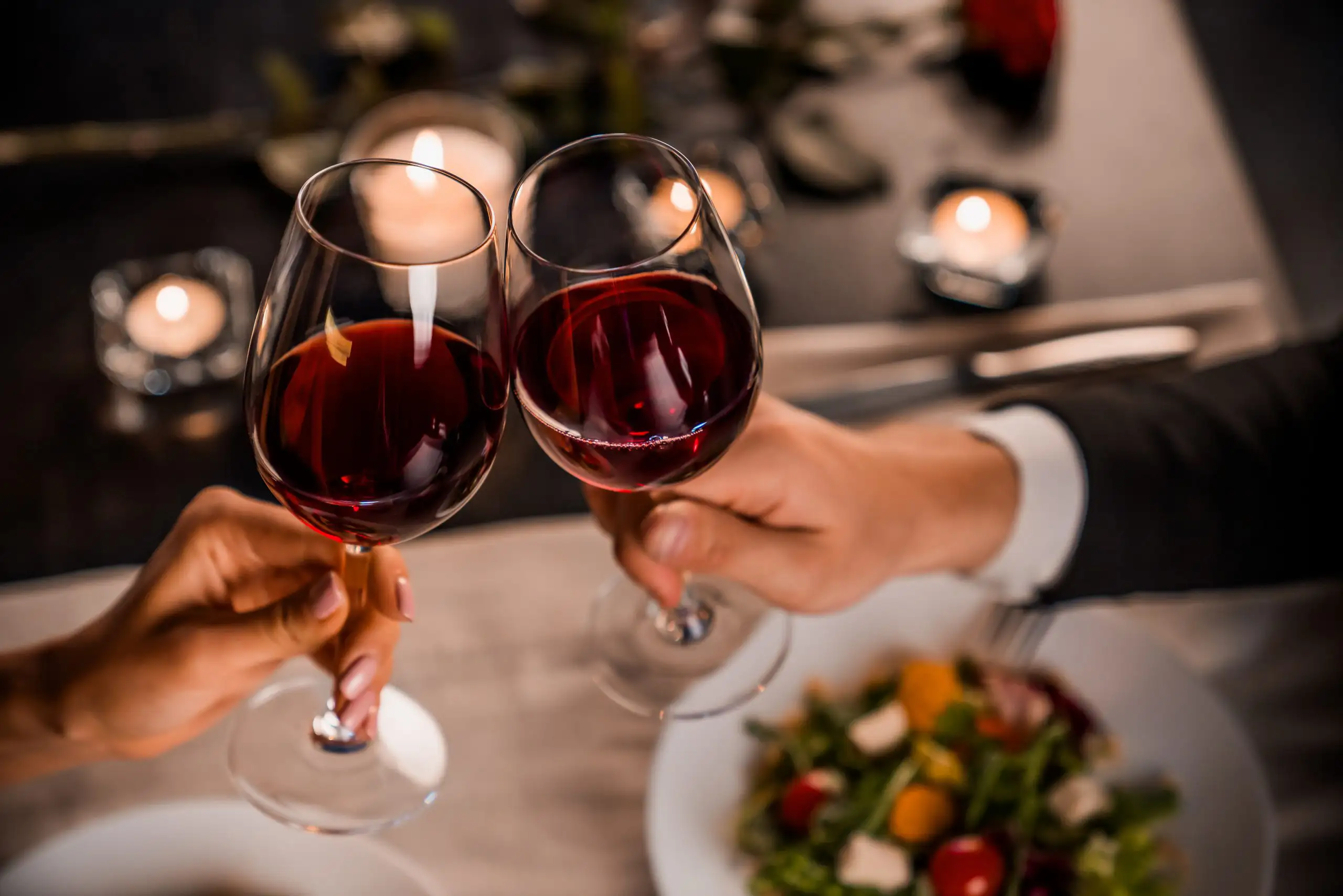 A bottle of red wine and a full wine glass on a table decorated with pink rose petals and red roses.
