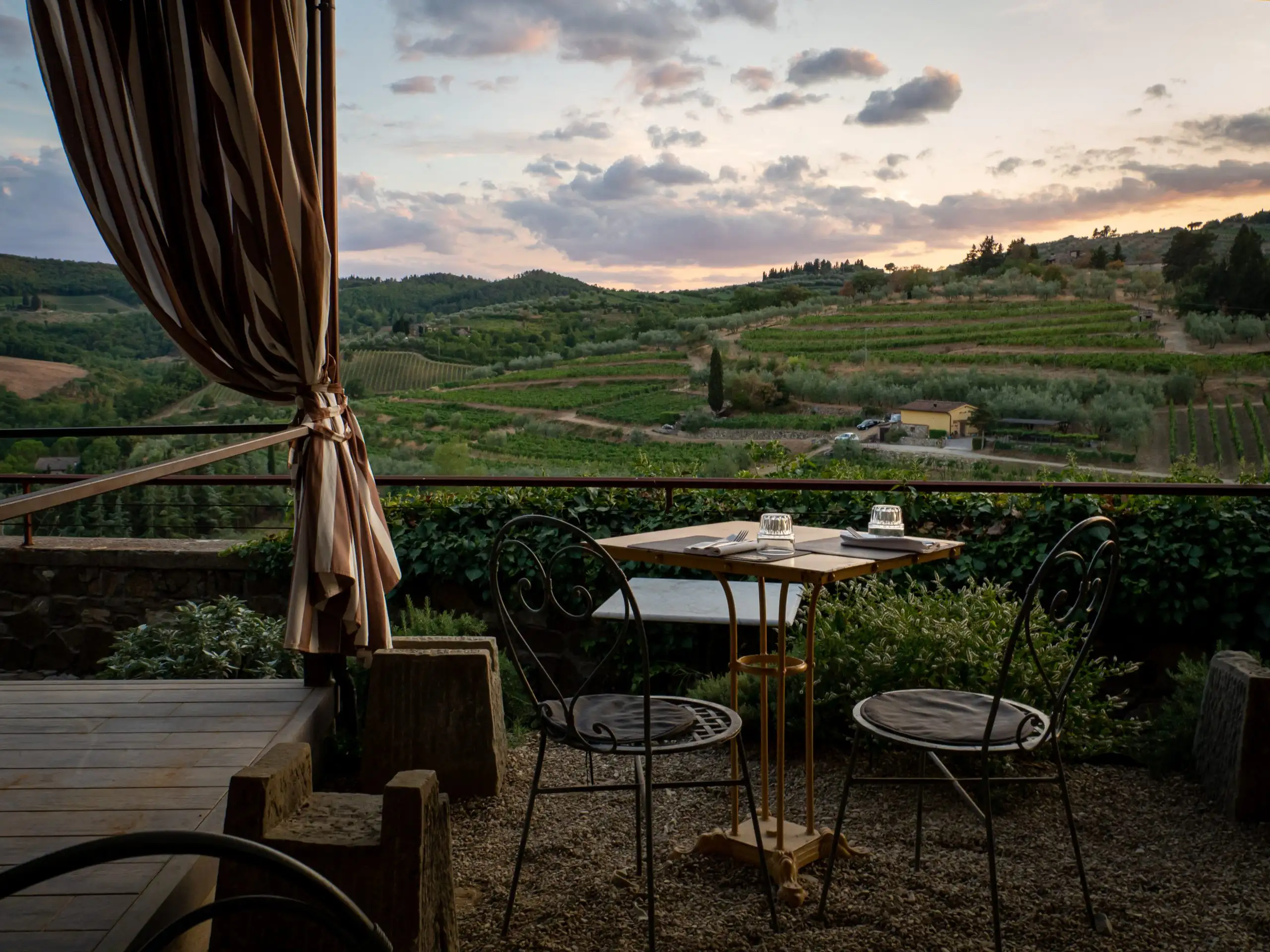 Outdoor restaurant patio with a small table and chairs overlooking terraced vineyards at sunset.